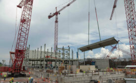 A large red crane lifts a gray rectangular slab with workers wearing yellow vests below, a cloudy sky in the background