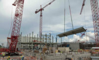 A large red crane lifts a gray rectangular slab with workers wearing yellow vests below, a cloudy sky in the background