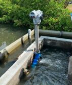 A corner of a stormwater pond is shown, with a valve standing vertically atop a metal pole. A river and bushes lie behind it