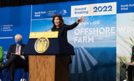 NY Governor Kathy Hochul gestures with her hand as she speaks behind a dais. In the background a large banner notes a 2022 groundbreaking for South Fork Wind Farm, "New York's First Offshore Farm." 