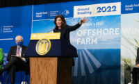 NY Governor Kathy Hochul gestures with her hand as she speaks behind a dais. In the background a large banner notes a 2022 groundbreaking for South Fork Wind Farm, "New York's First Offshore Farm." 