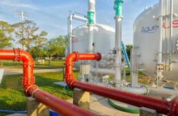 In this outdoor scene, orange red pipes shown in the foreground connect to two white water storage tanks. Trees and grass are seen in the background amidst a blue sky.  