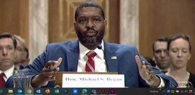 EPA Administrator Michael Regan wears a white shirt, red tie and blue jacket, talking in front of a mic at a hearing