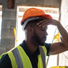 A construction worker wearing an orange hardhat and yellow vest puts a hand on their forehead, elbow resting again a wall