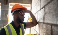 A construction worker wearing an orange hardhat and yellow vest puts a hand on their forehead, elbow resting again a wall