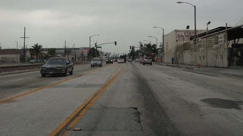 Cars drive along a road in South Los Angeles with visible bumps, under a gray-blue sky