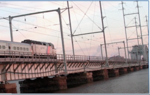 A New Jersey Transit train crosses over a bridge, with a pink and blue sky in the background.