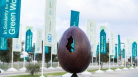 A sculpture of an egg with a crack is shown in a courtyard with green banners displaying COP29 logos