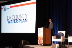 Speaker wearing a suit stands behind a podium on a stage. Near him, a projector reads, "LA County Water Plan"