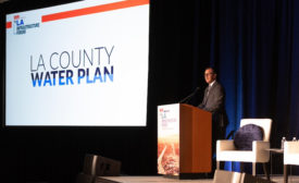 Speaker wearing a suit stands behind a podium on a stage. Near him, a projector reads, "LA County Water Plan"