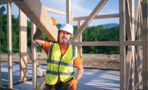 A construction worker wearing an orange shirt and gloves, white hardhat and yellow vest holds up a wooden beam, with the frame of a structure in the background. 